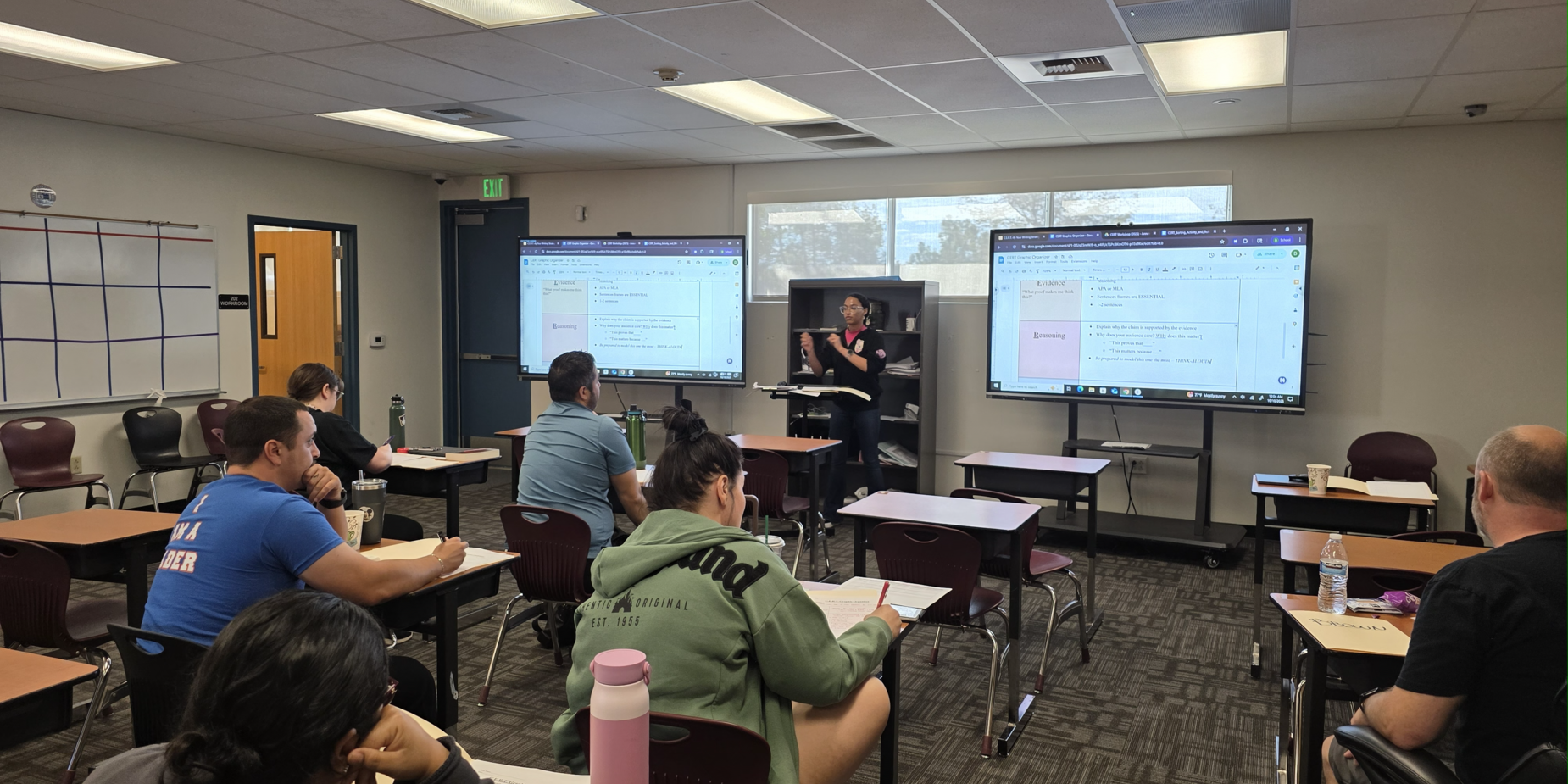 Classroom with students seated at desks, instructor presenting on dual screens.