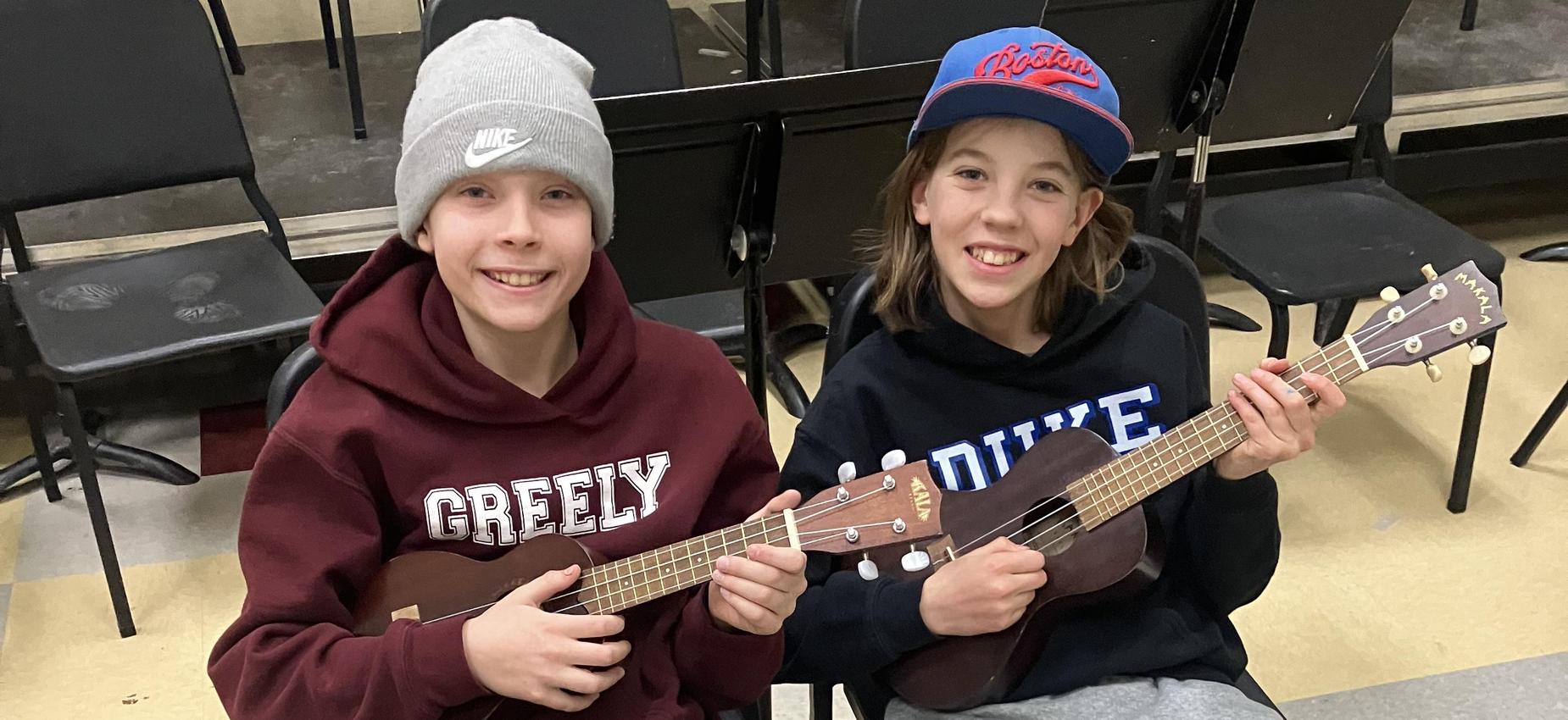 Two children seated in chairs, playing ukuleles in a classroom setting.