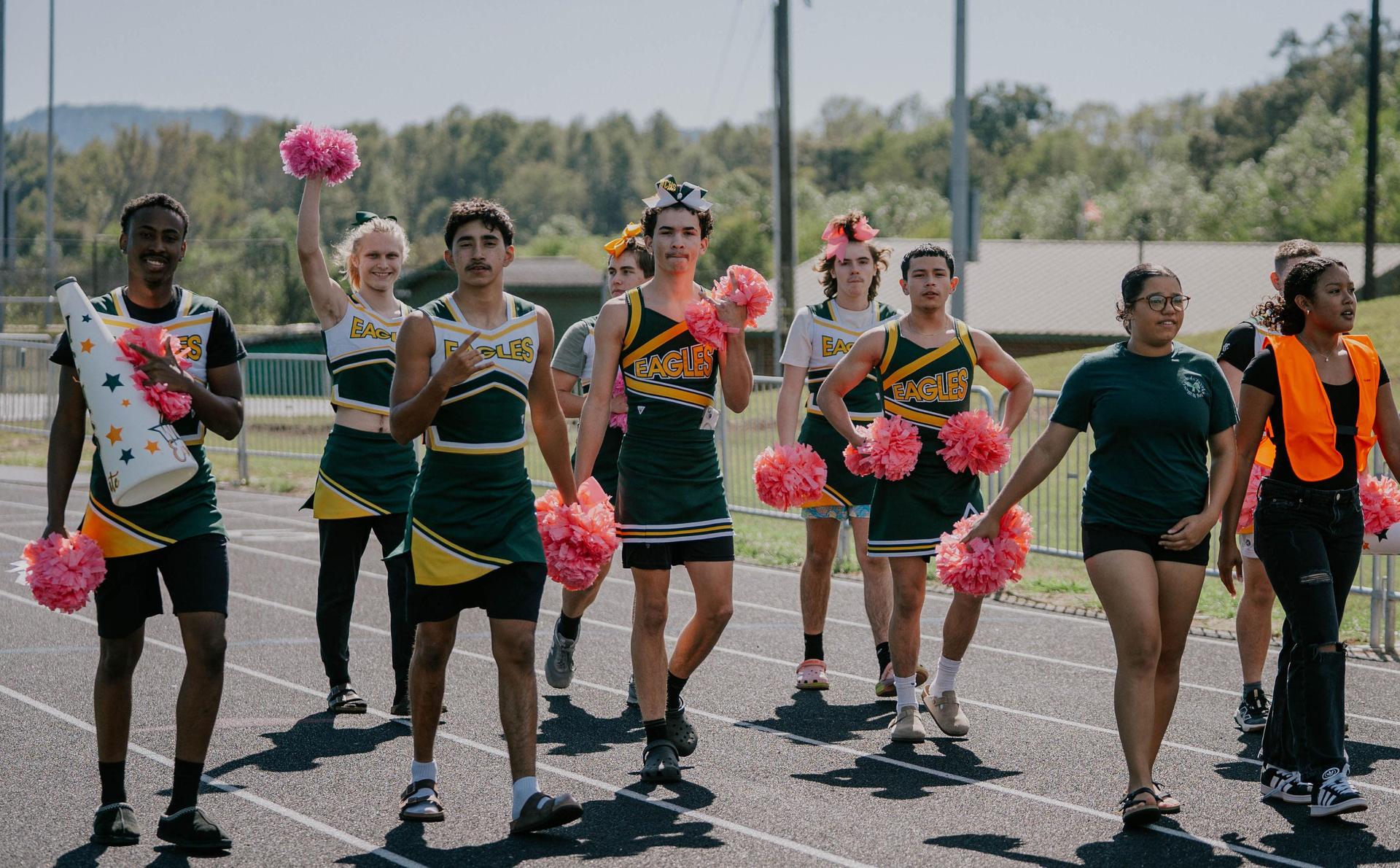 Cheerleaders in green and yellow uniforms march with pom-poms on a track.