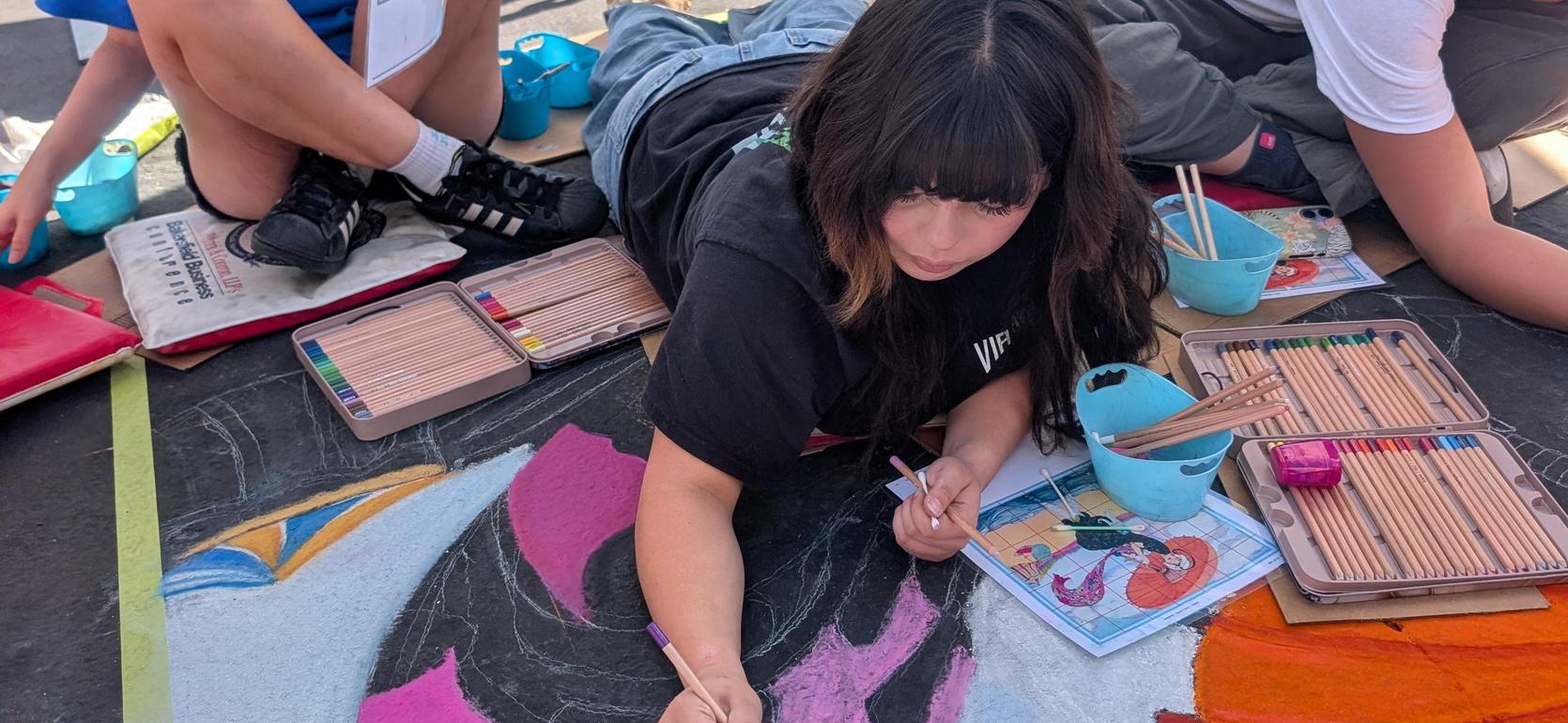 A person lying on their stomach adds color to a chalk mural, surrounded by art supplies.