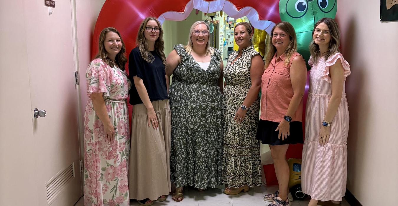 Six women pose together in front of a colorful inflatable arch in a hallway.