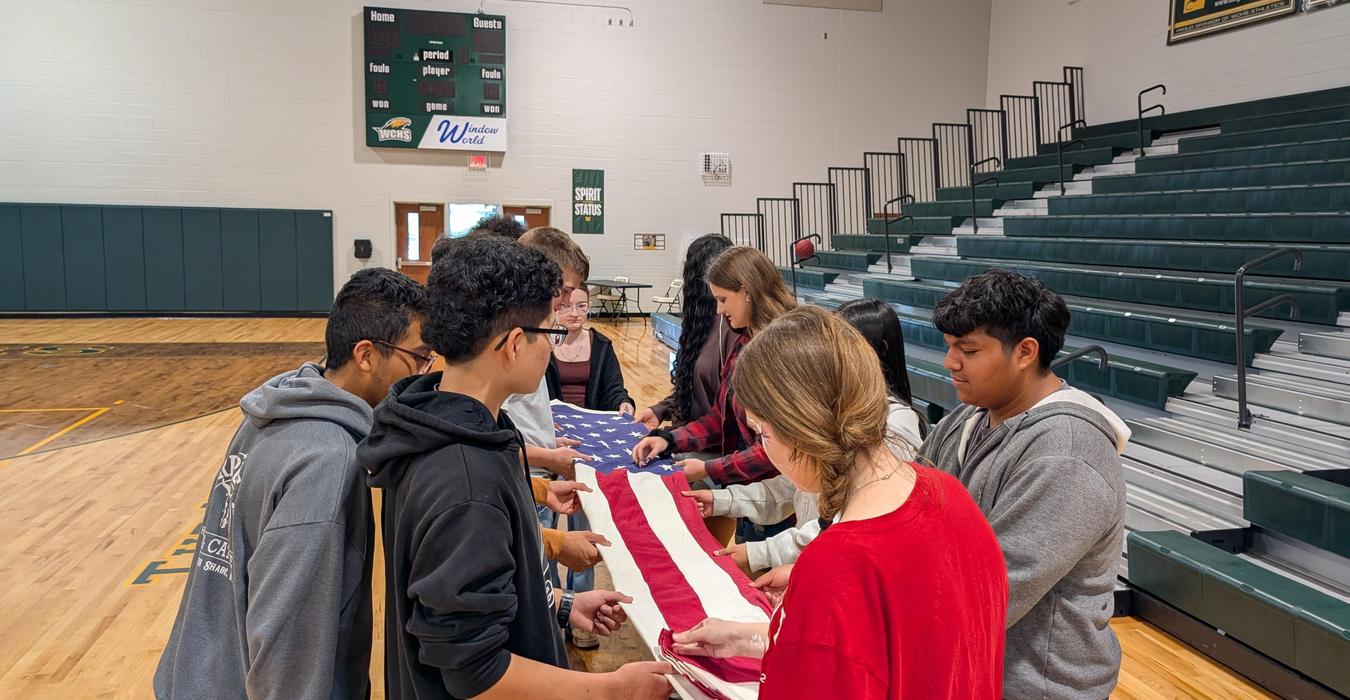 Students collaboratively hold and arrange a large American flag in a gym.