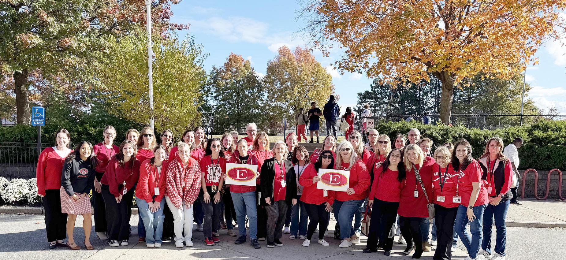 A group of approximately 40 people wearing red shirts, gathered outdoors with autumn foliage.