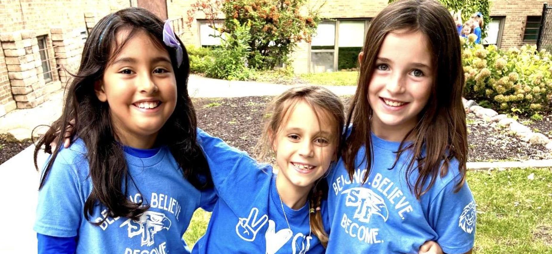 Three girls in blue shirts smiling and posing together outdoors.