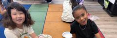 Two children sitting on multi-colored carpet tiles, smiling at the camera.