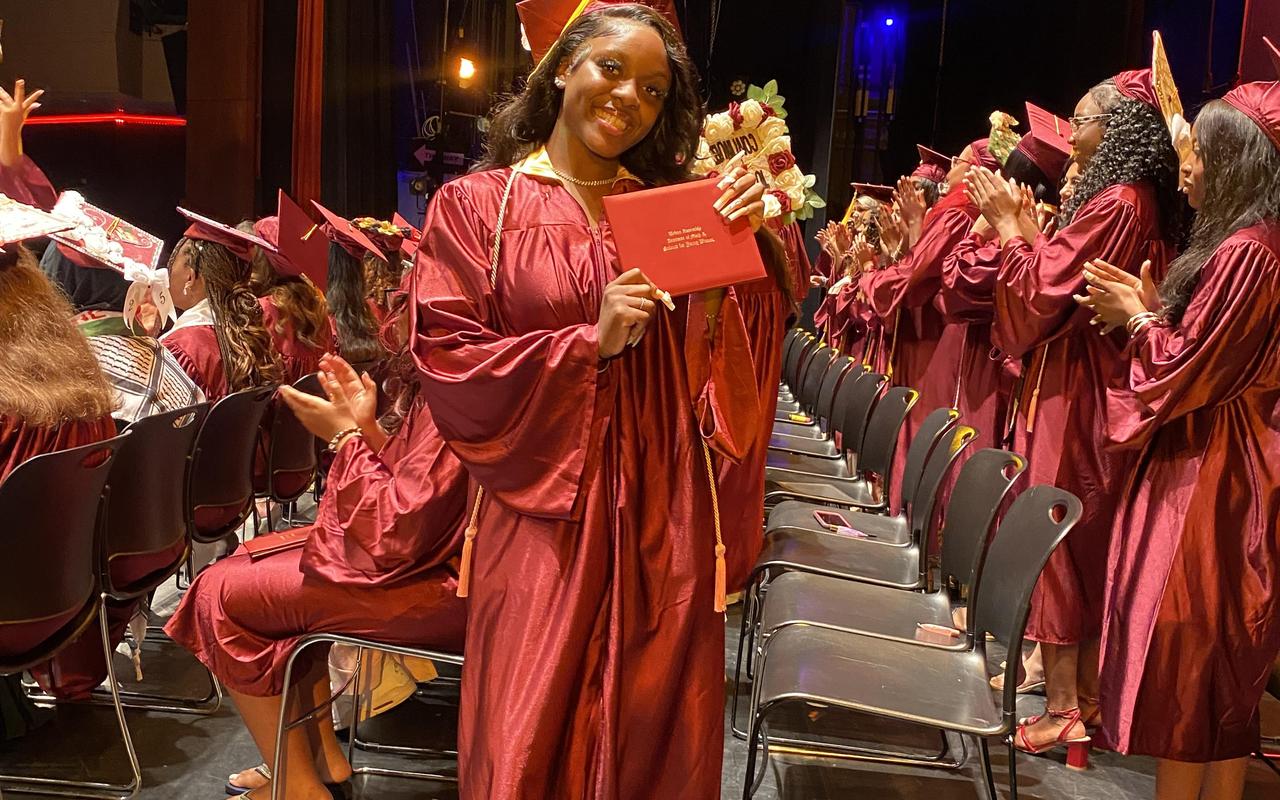A graduate in a maroon gown holds a diploma, surrounded by peers in a celebratory atmosphere.