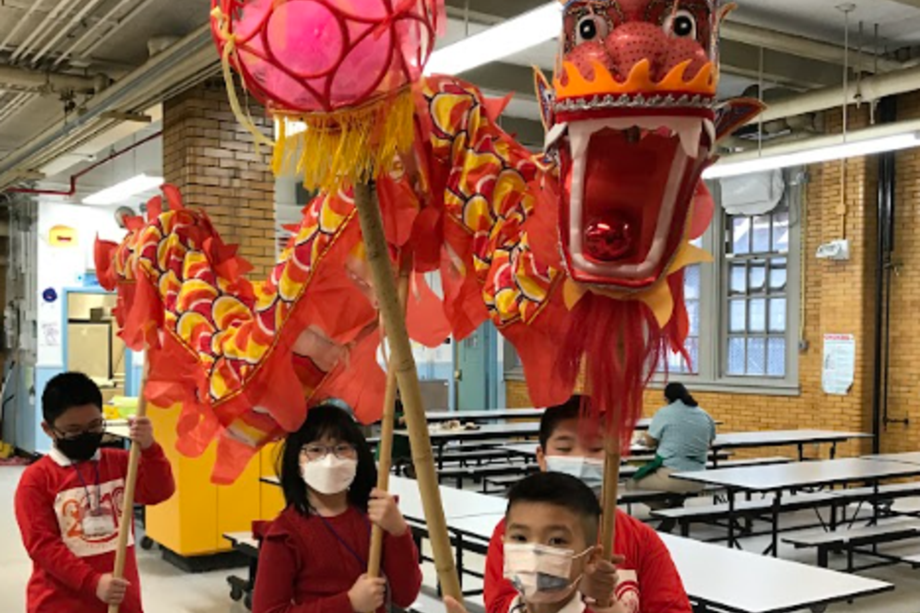 Children in red shirts holding a dragon lantern and a dragon puppet in a school cafeteria.