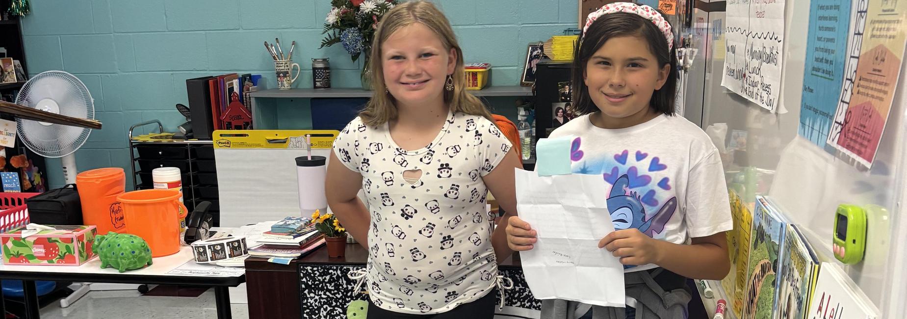 Two girls holding a paper and standing in a colorful classroom setting.