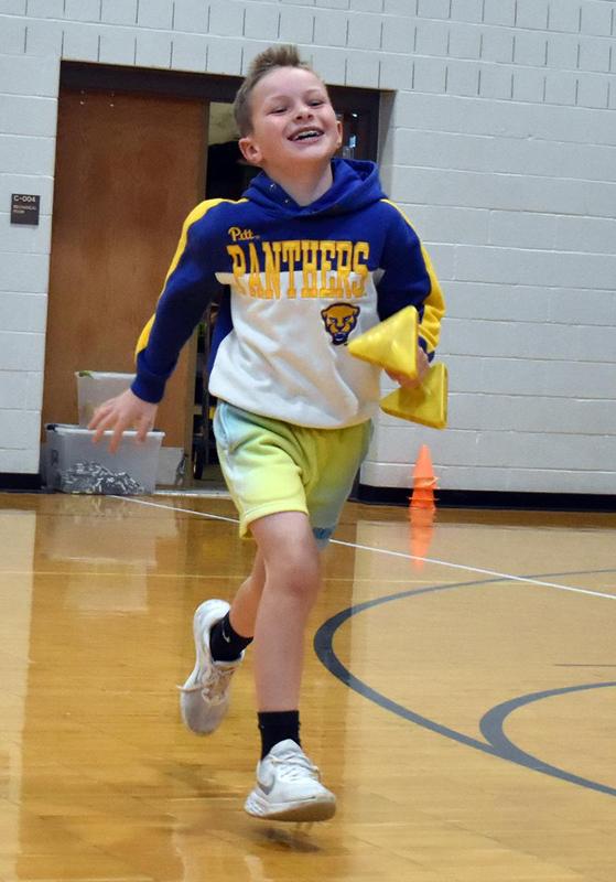 A young boy jogs in a gym holding yellow objects, smiling and wearing a colorful hoodie.