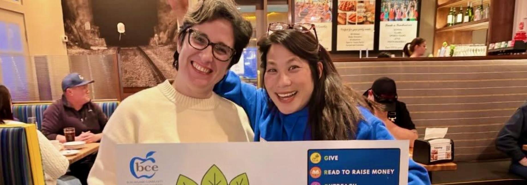 Two smiling women holding a sign promoting a fundraising event at a restaurant.