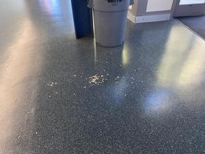 Dark speckled commercial flooring with scattered crumbs or food debris near a gray Brute trash can and blue recycling bin. A door mat and white cabinet base are visible in the background.
