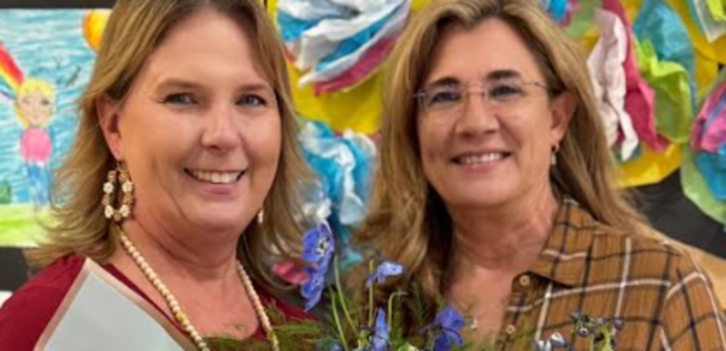 Two smiling women holding a bouquet of flowers and a bottle.