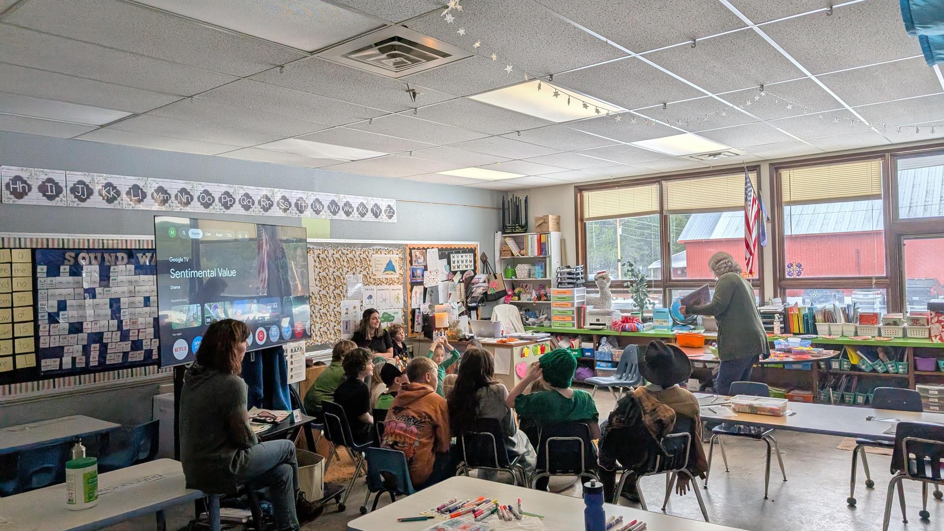 Classroom scene with students, a teacher, and a projector displaying content.
