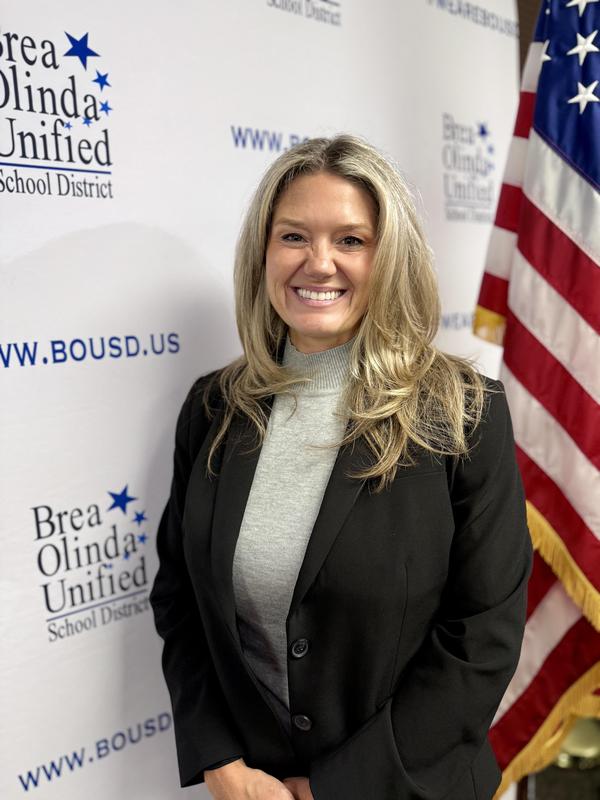Natalie Johnson wearing a black suit and gray turtleneck stands in front of a BOUSD backdrop. American flag to the side of her