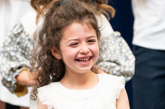 Smiling girl with curly hair in a light dress, surrounded by people.