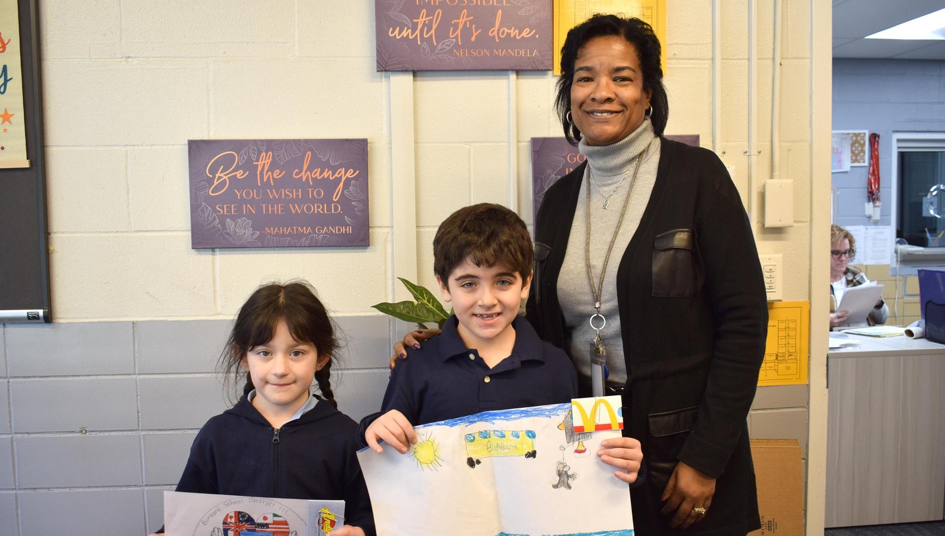 A teacher posing with two children showing their artwork that includes a beach scene.