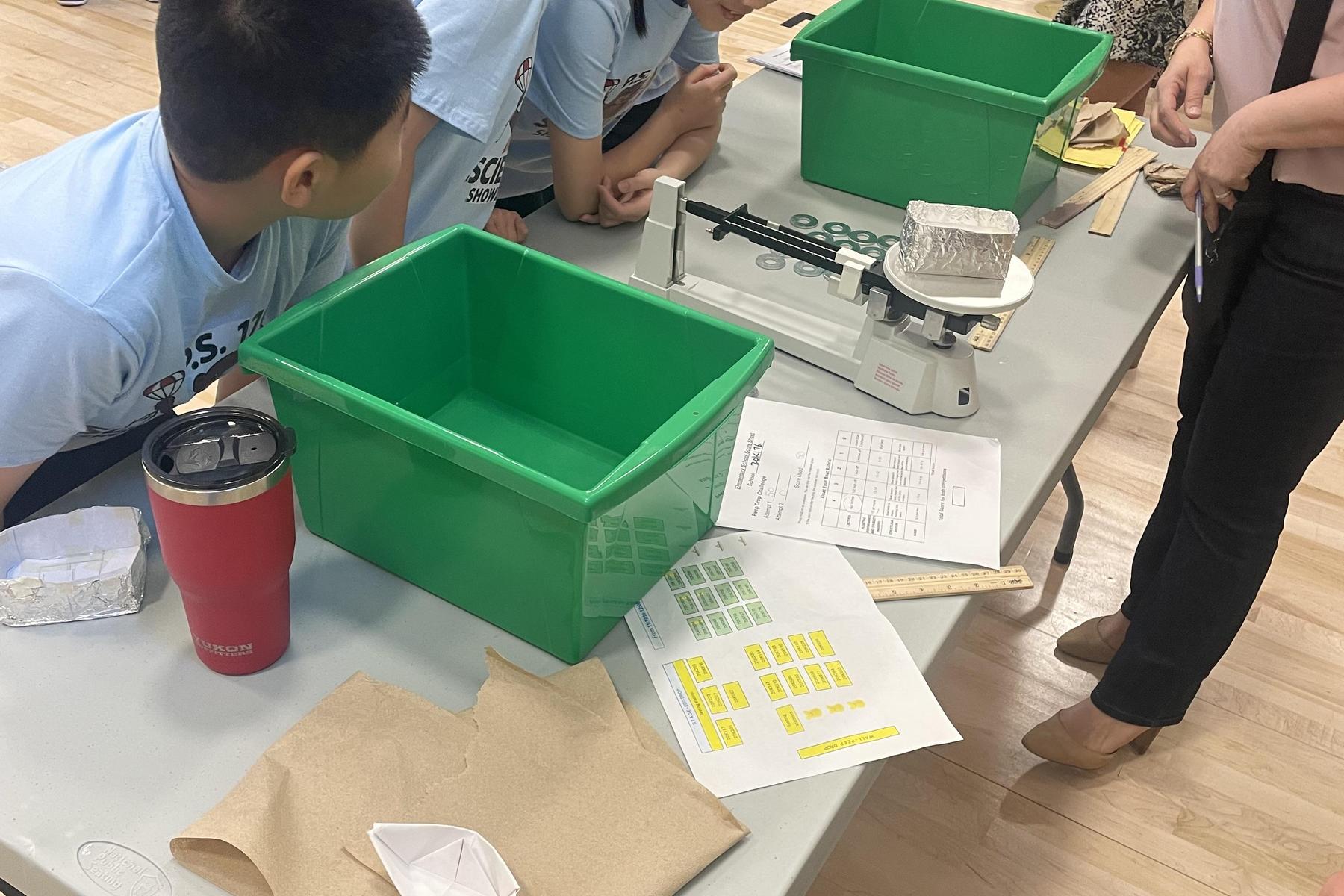 Kids gather around a table with green containers, materials, and a scale for an activity.