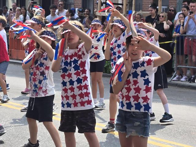 Children in patriotic outfits dance while waving ribbons at a parade.