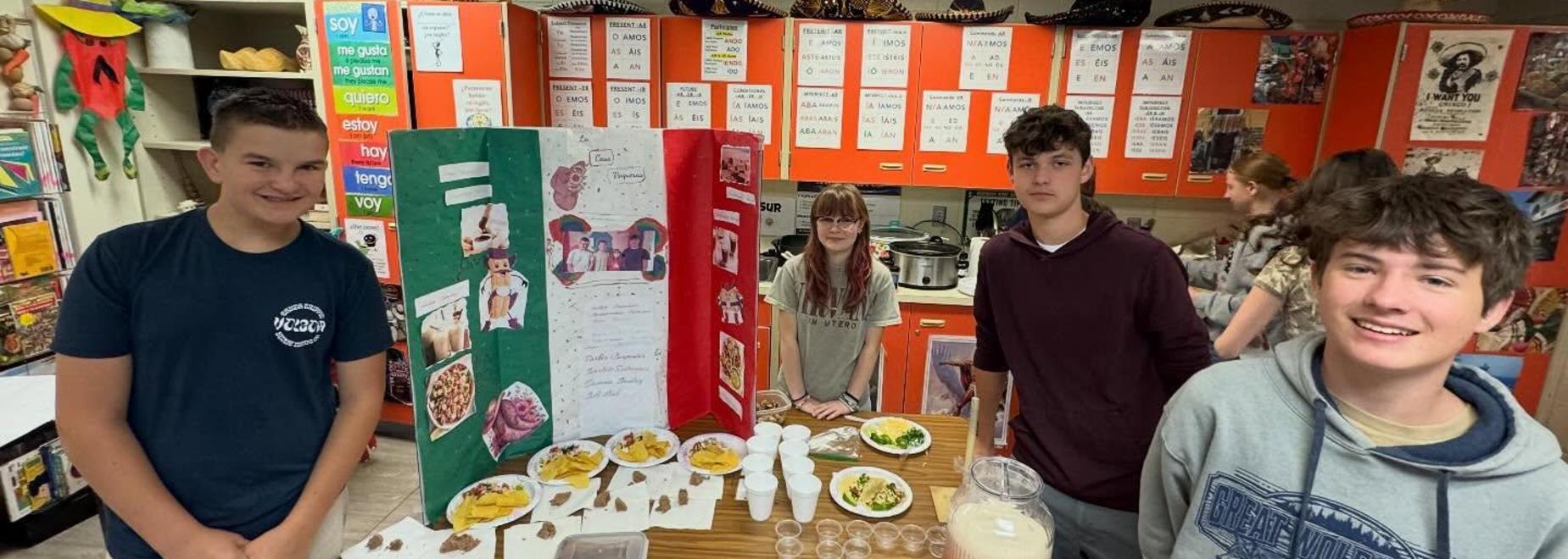 Four students present a food display with a tri-fold poster in a colorful classroom.