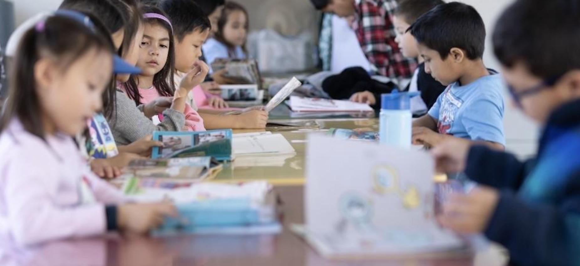 Children reading books at a table in a classroom setting.