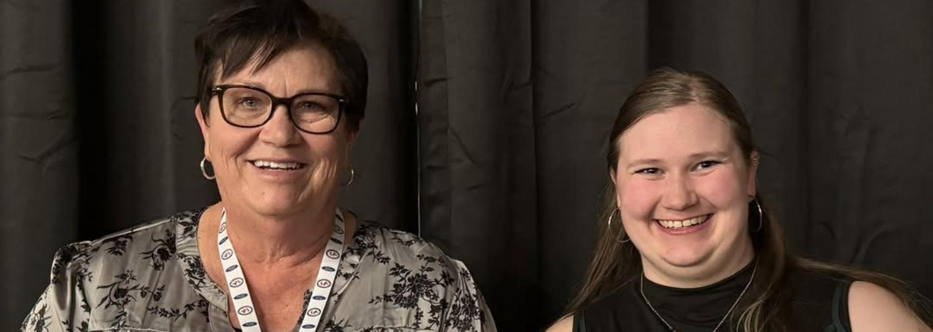 Two women smiling, each holding a plaque, standing in front of a dark curtain.