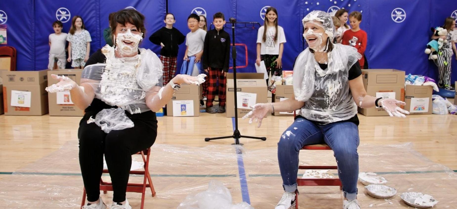 Two women covered in whipped cream smile while sitting in chairs, with kids in the background.