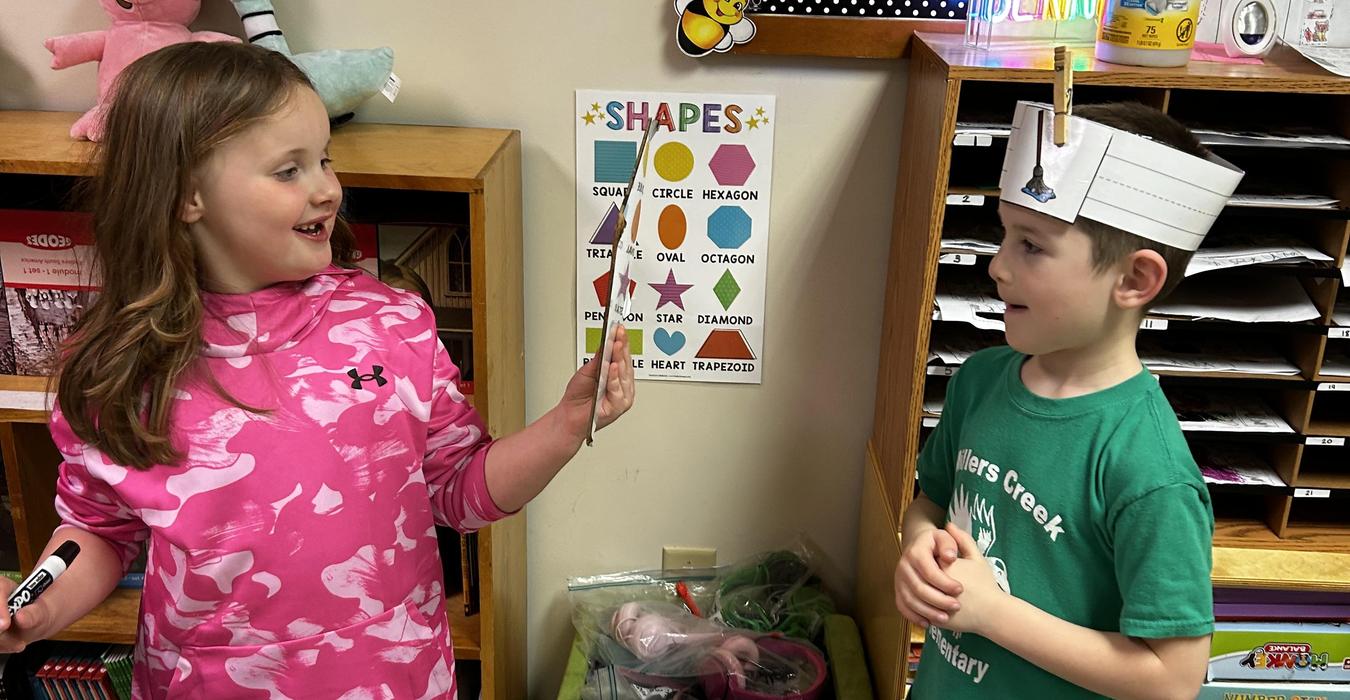 Two children interacting, one holding a marker and the other wearing a hat with shapes.