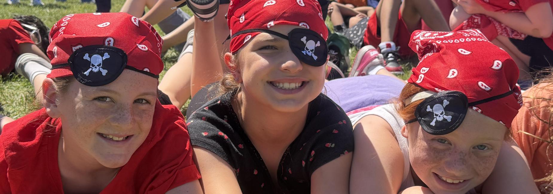 Three girls wearing pirate bandanas and eyepatches, smiling in a grassy area.