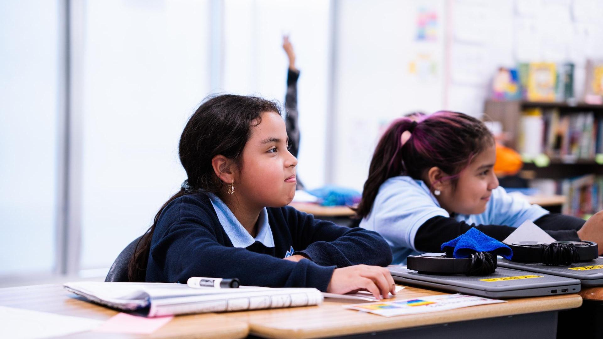 Two girls in a classroom, one raising a hand and the other focused.