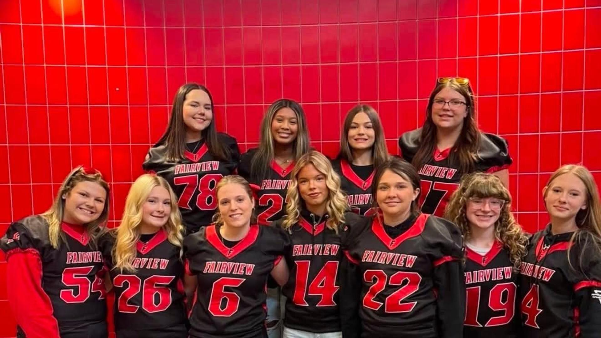 Group of girls in football jerseys posing together in front of a red wall.