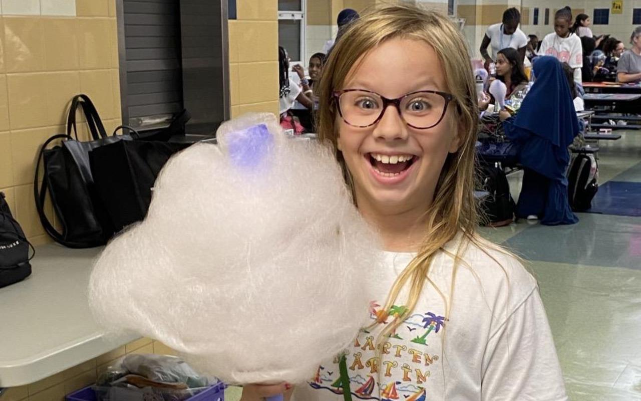 Child smiling while holding a large cotton candy in a school setting.