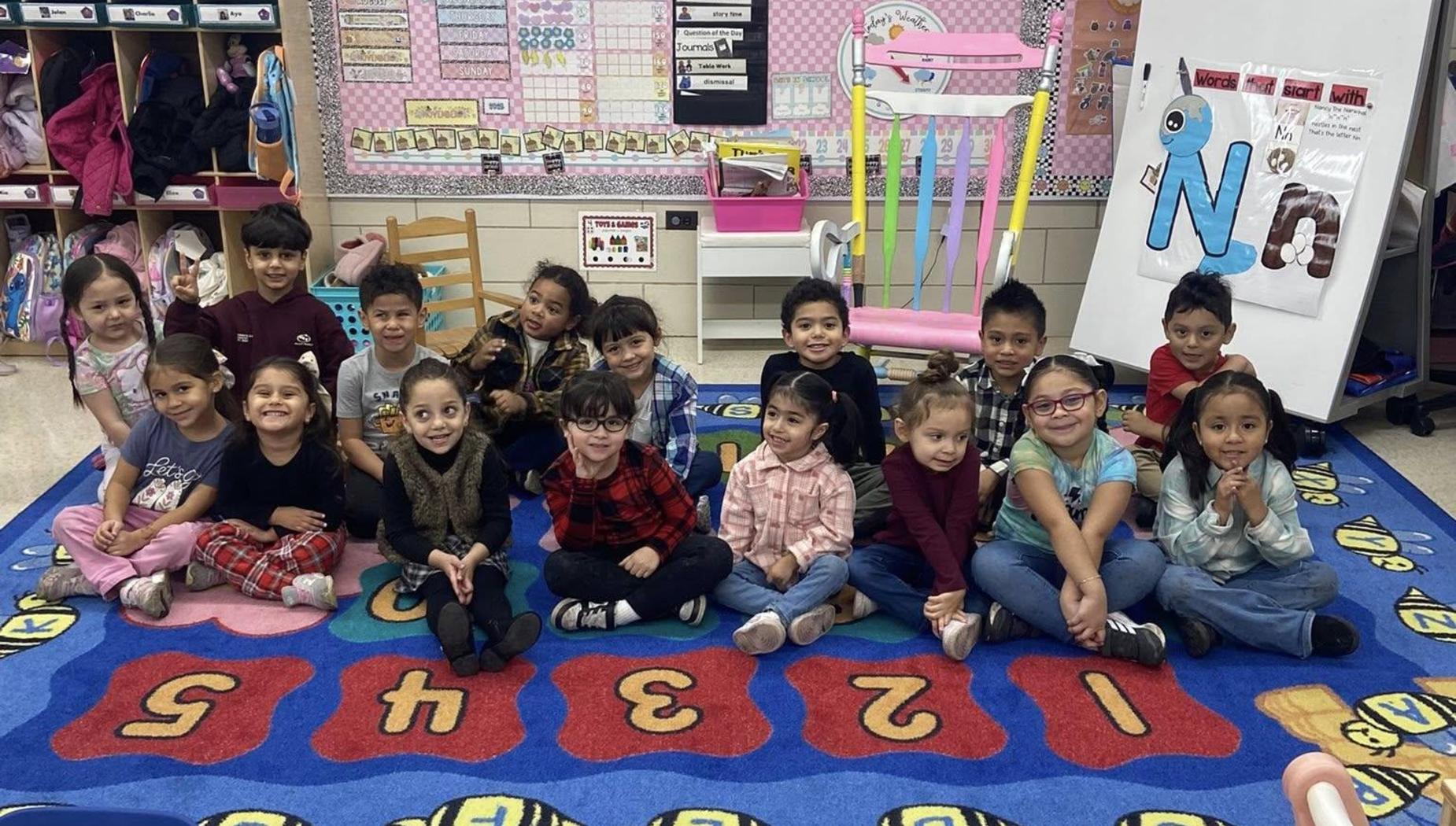 A large group of children sitting on a rug, smiling and posing for the photo in a classroom.