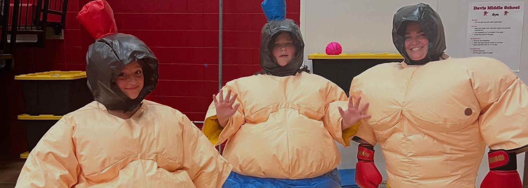 Three individuals stand together in sumo suits, smiling and waving.