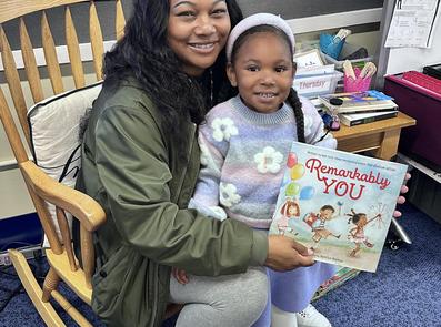 A woman and a girl smiling while holding a children's book in a classroom.