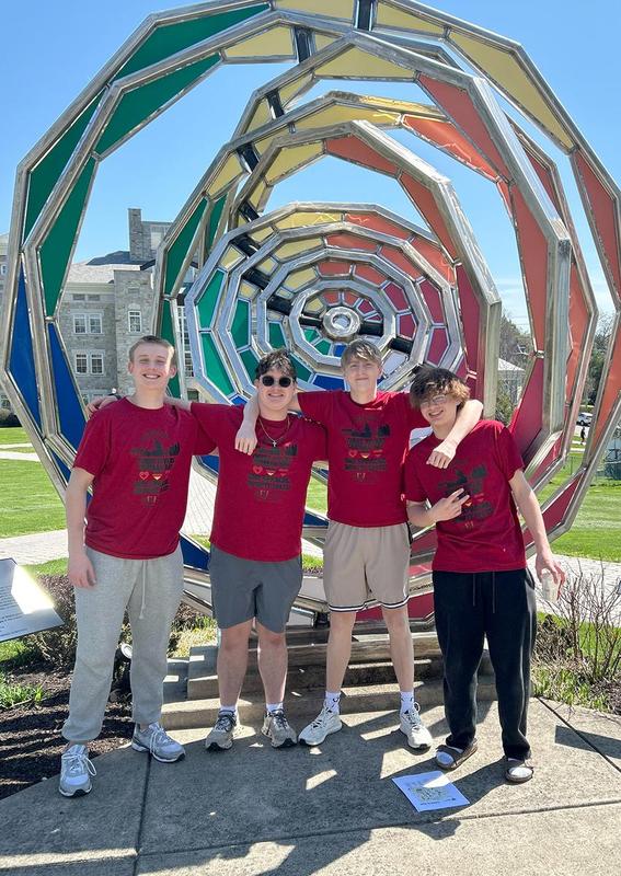 Four students in red shirts posing in front of a colorful spiral sculpture.