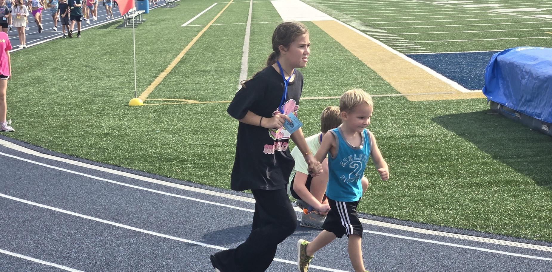 A young girl walking hand-in-hand with a child on a track, with others running in the background.