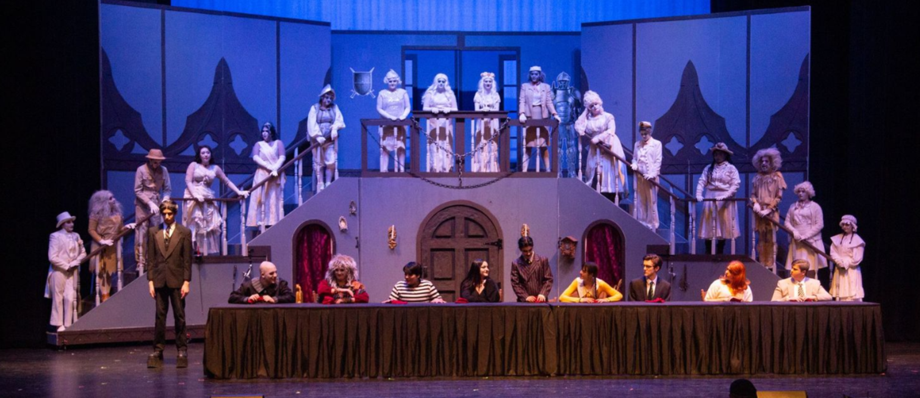Addams Family performance with ghostly characters up the stairs on stage and the family at a long table