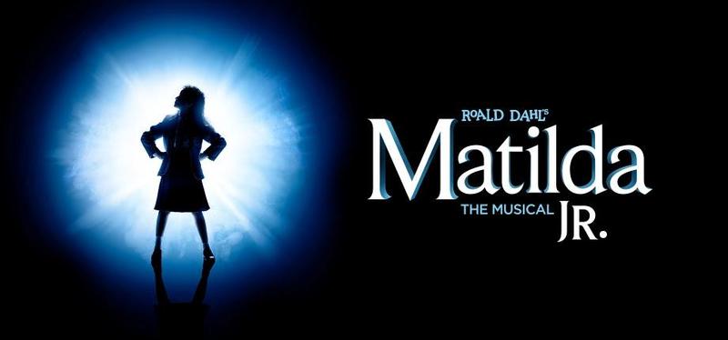 Matilda, the character from the musical, is standing in a spotlight on a stage.