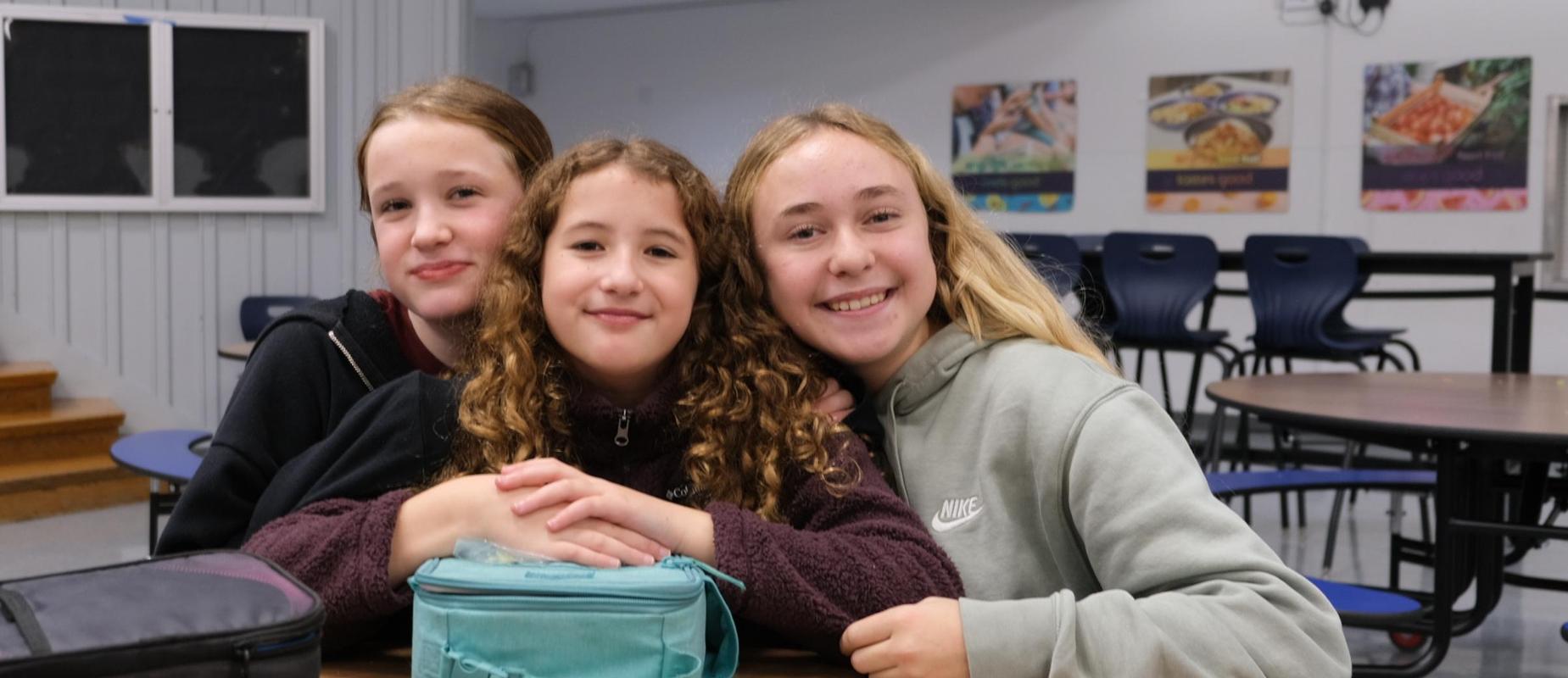 Three smiling girls sitting at a table in a cafeteria, with bags nearby.