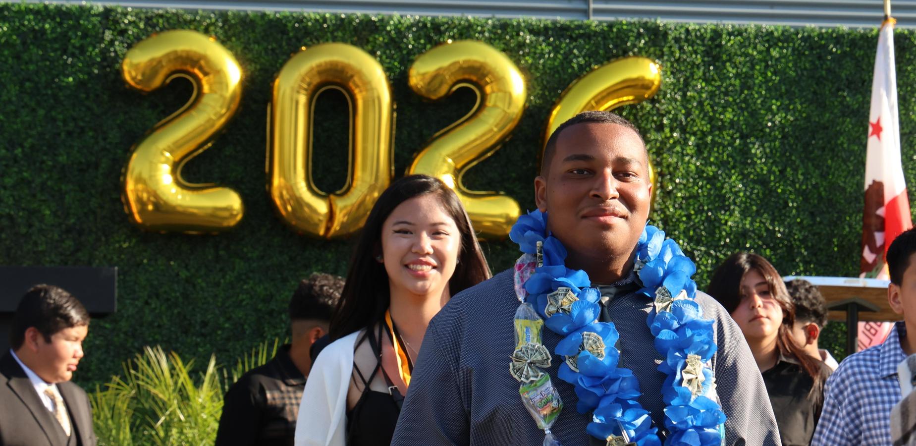 Students celebrating graduation with balloons and leis in front of a school building.