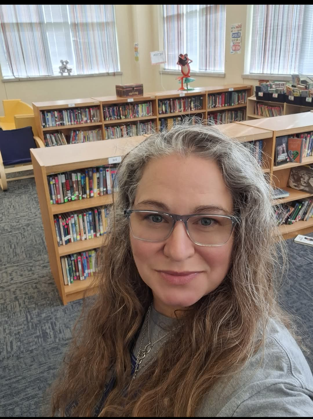 A woman with long hair and glasses smiling in a library filled with books.
