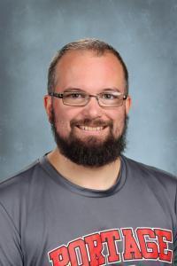 Smiling man with a beard wearing a Portage shirt against a blue background.
