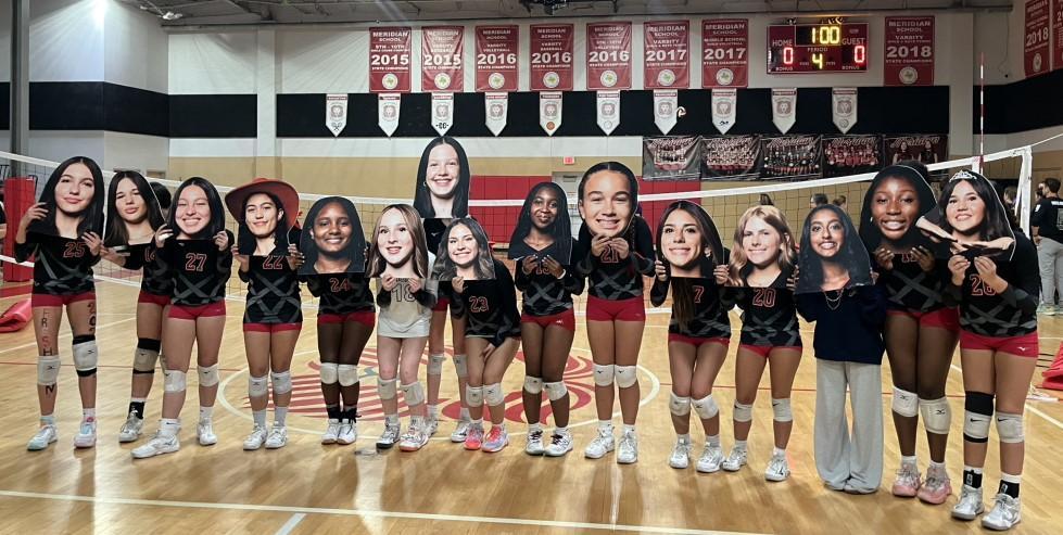 Girls' Varsity Volleyball team members pose with giant cut-out posters of their heads in the Mays Gym