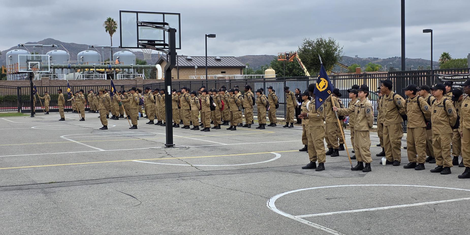 A large group of uniformed cadets lined up on a basketball court, with mountains in the background.
