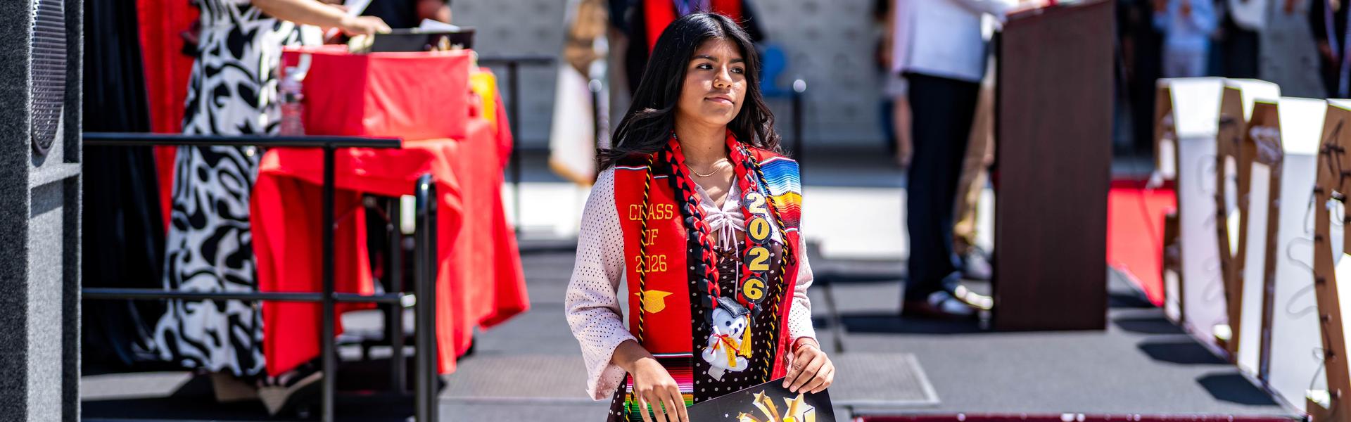 Graduate standing with a decorated poster and colorful sash at a ceremony.