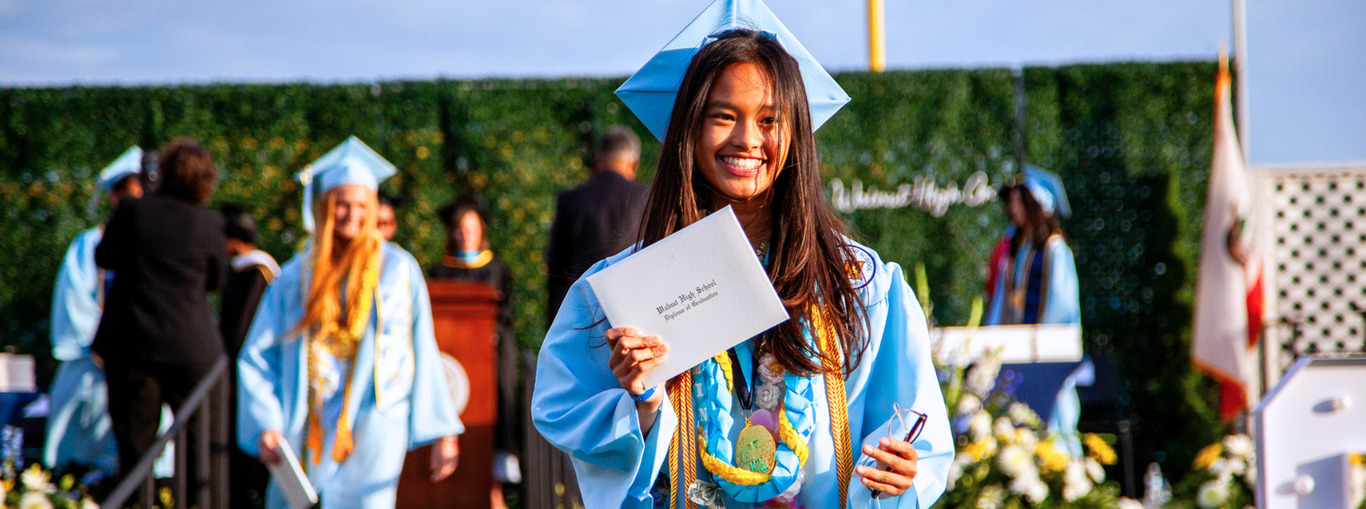 Graduate in blue gown holding a diploma with other graduates in the background.