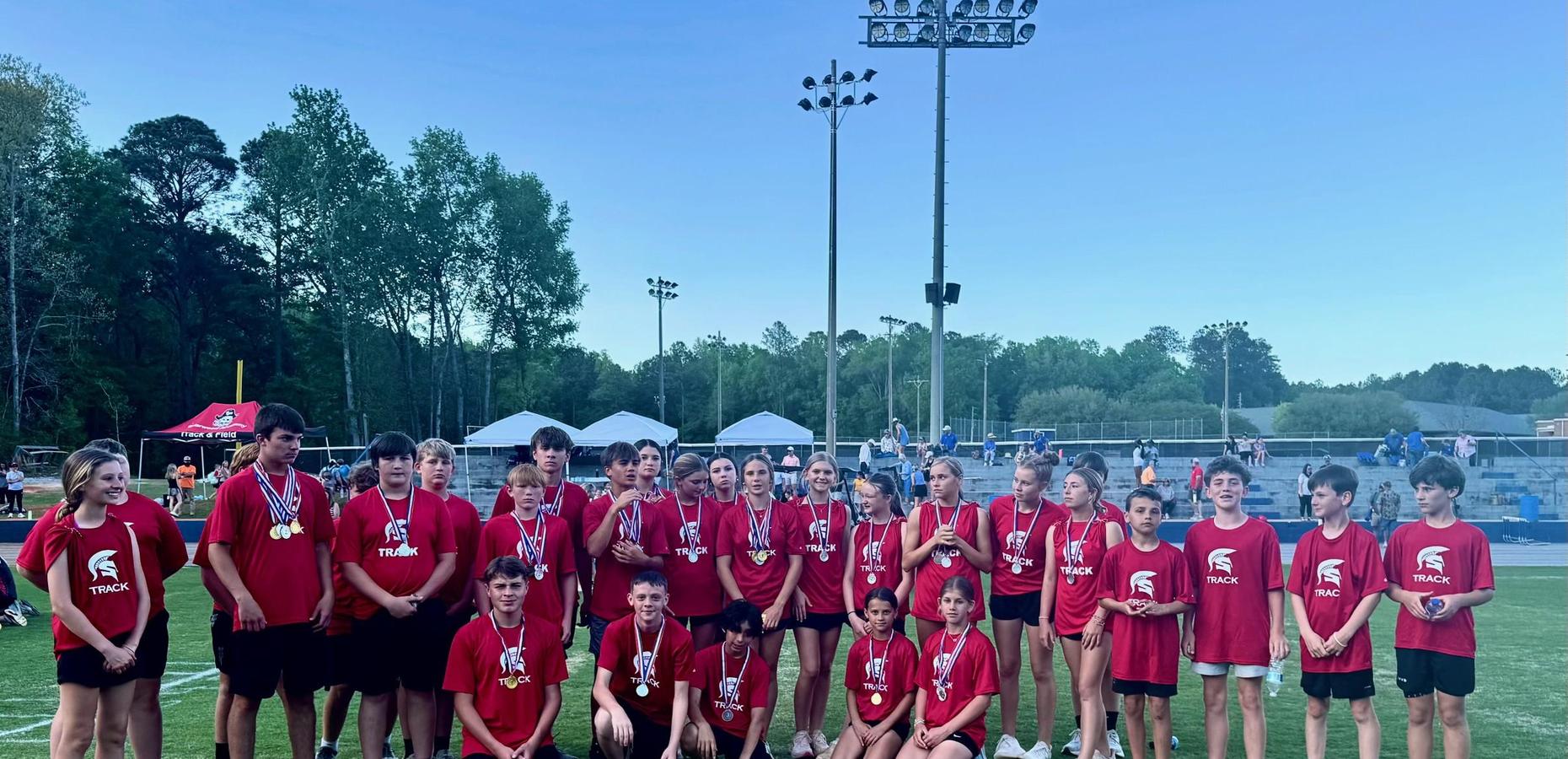 Team of young athletes in red shirts, displaying medals at a track event.