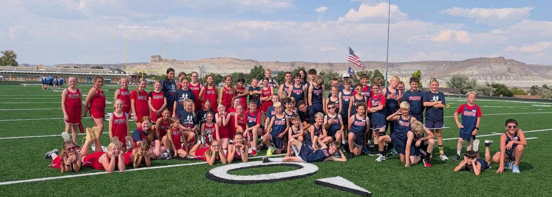 A large group of young athletes in red and blue uniforms posing on a sports field with mountains in the background.