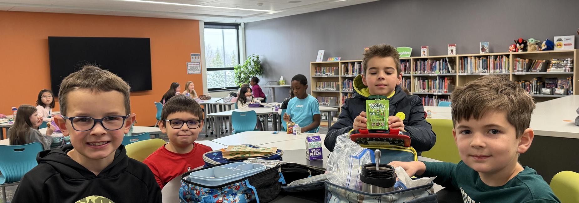 Four children at a classroom table with backpacks and lunchboxes, smiling and engaged.