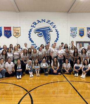 Group of people posing together in a gym with banners and a logo on the wall.